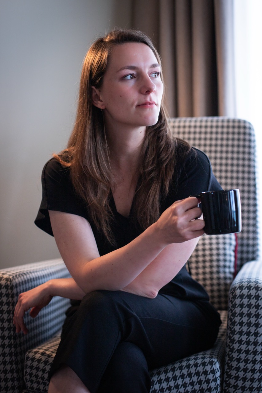 Young woman in black jumpsuit looking into the distance with a coffee cup.